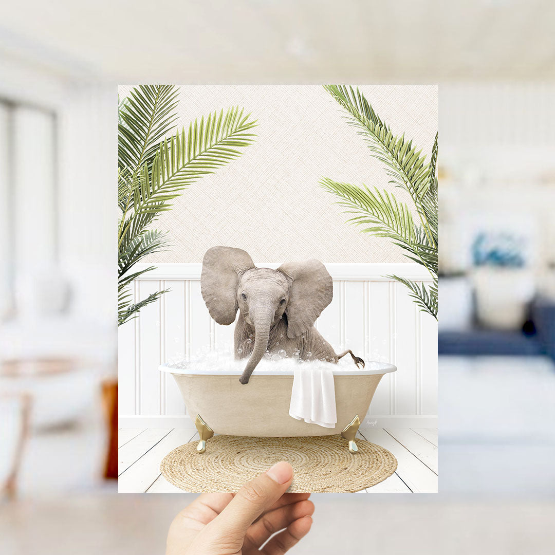 A hand holds up a framed image depicting a baby elephant sitting in a bathtub, surrounded by palm leaves.