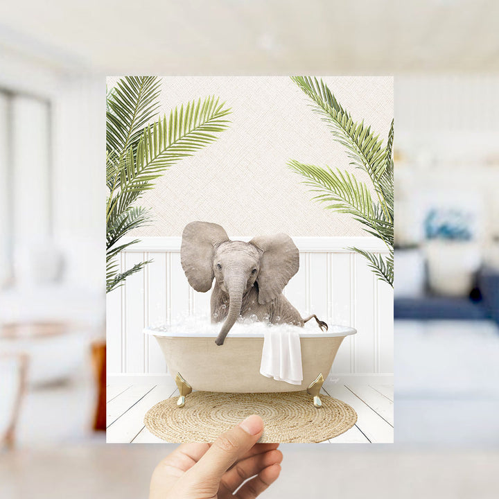 A hand holds up a framed image depicting a baby elephant sitting in a bathtub, surrounded by palm leaves.