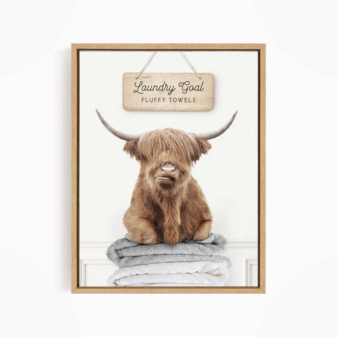A framed image of a brown cow with long horns sitting on a stack of fluffy towels, with a sign above that says "Laundry Goal Fluffy Towels".