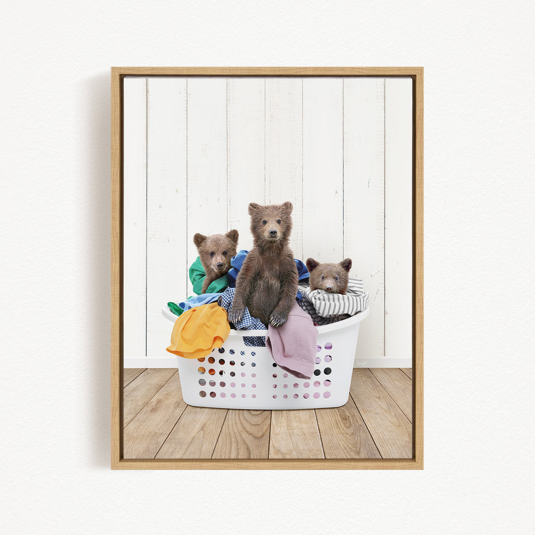 A framed image showing three brown bears sitting in a white laundry basket filled with clothes.
