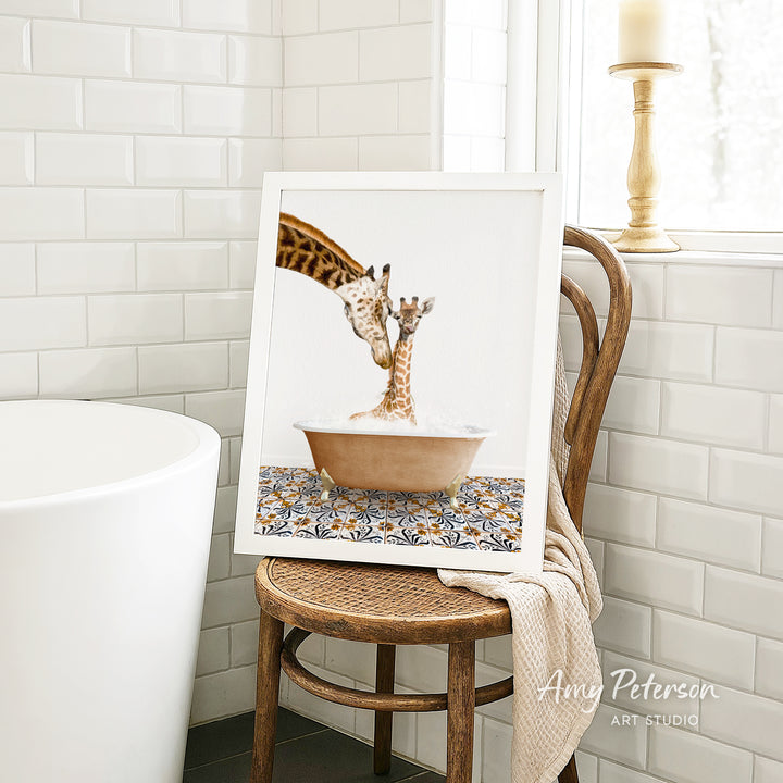 A framed image of a giraffe and a baby giraffe sitting in a bathtub, with a towel draped over the back of a wooden chair in a bathroom setting.