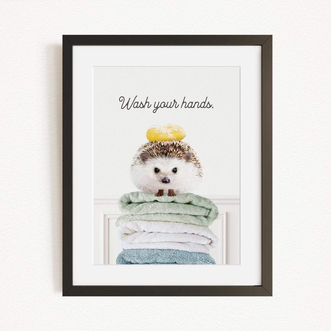 A framed image of a hedgehog sitting atop a stack of folded towels, with the text "Wash your hands" written above it.