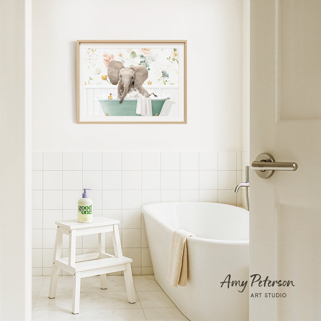 A framed painting of two elephants sitting in a bathtub is hanging on a white tiled wall above a white bathtub in a bathroom.