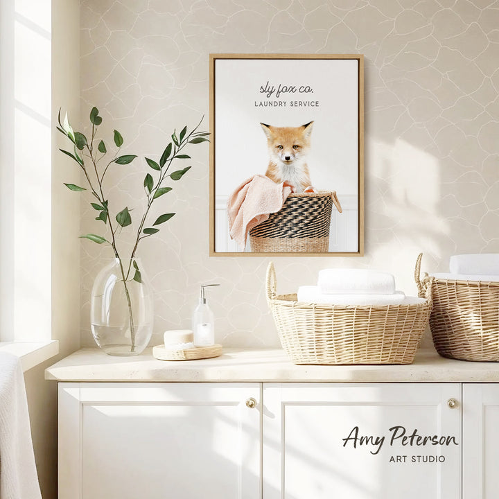 a framed photo of a cat sitting in a basket, with the text "Sly Fox Co. Laundry Service" above it. The photo is displayed on a wall above a white cabinet with baskets and a vase of leaves.
