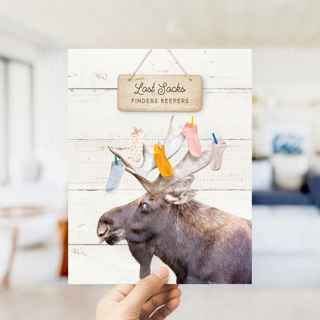 A hand is holding a card with a moose's head and antlers, with a sign that says "Lost Socks Finders Keepers" above it.