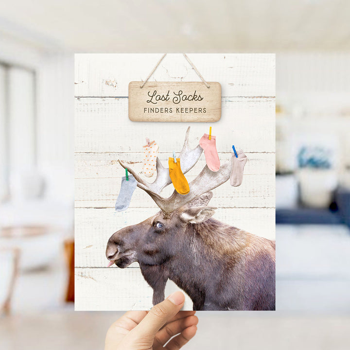 A hand is holding a card with a moose's head and antlers, with a sign that says "Lost Socks Finders Keepers" above it.