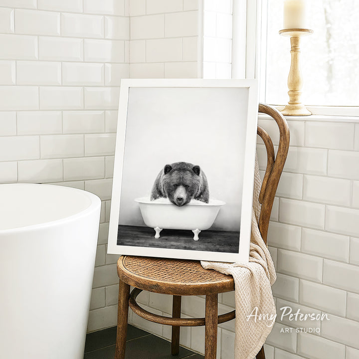 A framed black and white photograph of a bear sitting in a bathtub is displayed on a wooden chair in a bathroom setting.