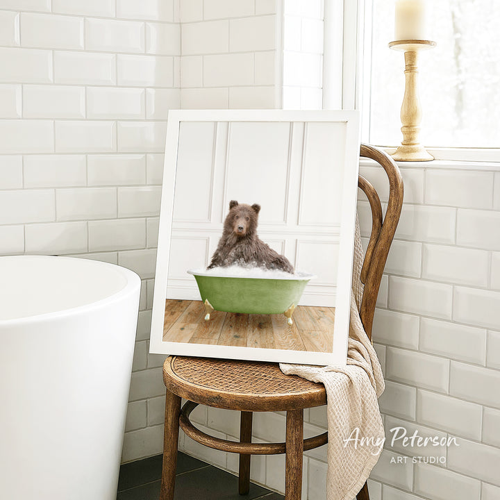 A framed image of a brown bear sitting in a green bathtub, with a towel draped over the back of a wooden chair in a bathroom setting.