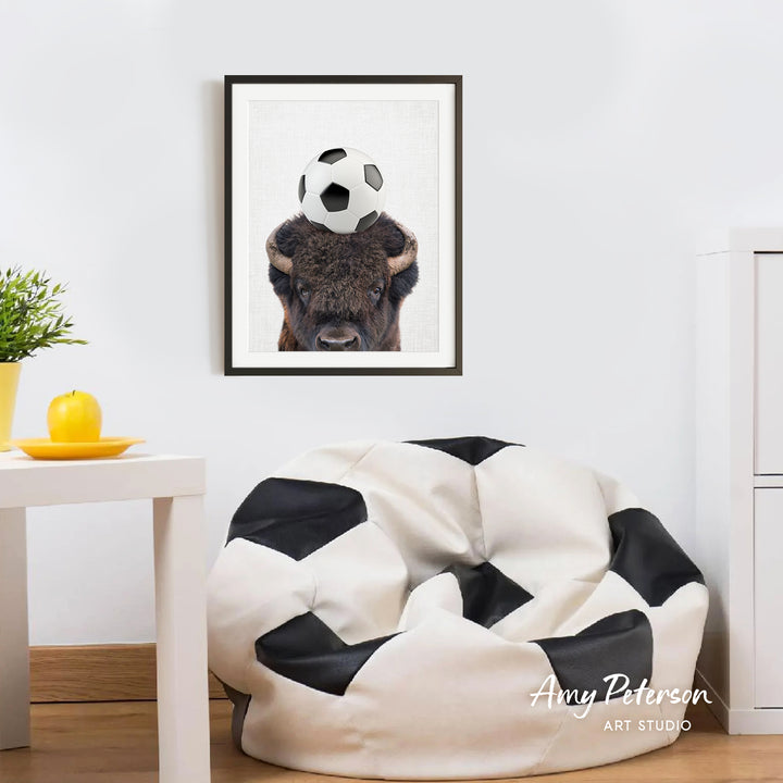 a framed picture of a bison with a soccer ball on its head, hanging on a white wall above a white table with a yellow plate on it.
