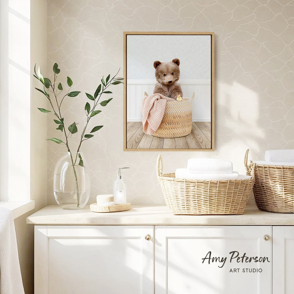 a cozy bathroom scene with a framed photo of a cute bear sitting in a basket, a vase with green leaves, and baskets on a white countertop.