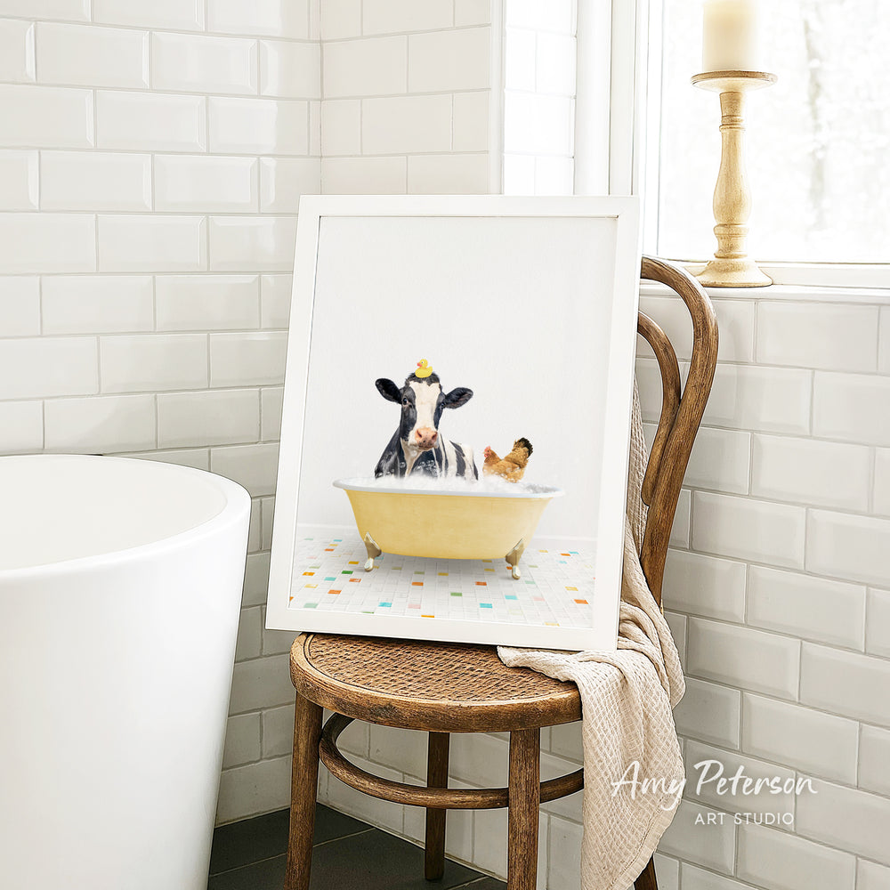 A framed image of a cow sitting in a bathtub, with a chicken nearby, is displayed on a wooden chair in a bathroom setting.