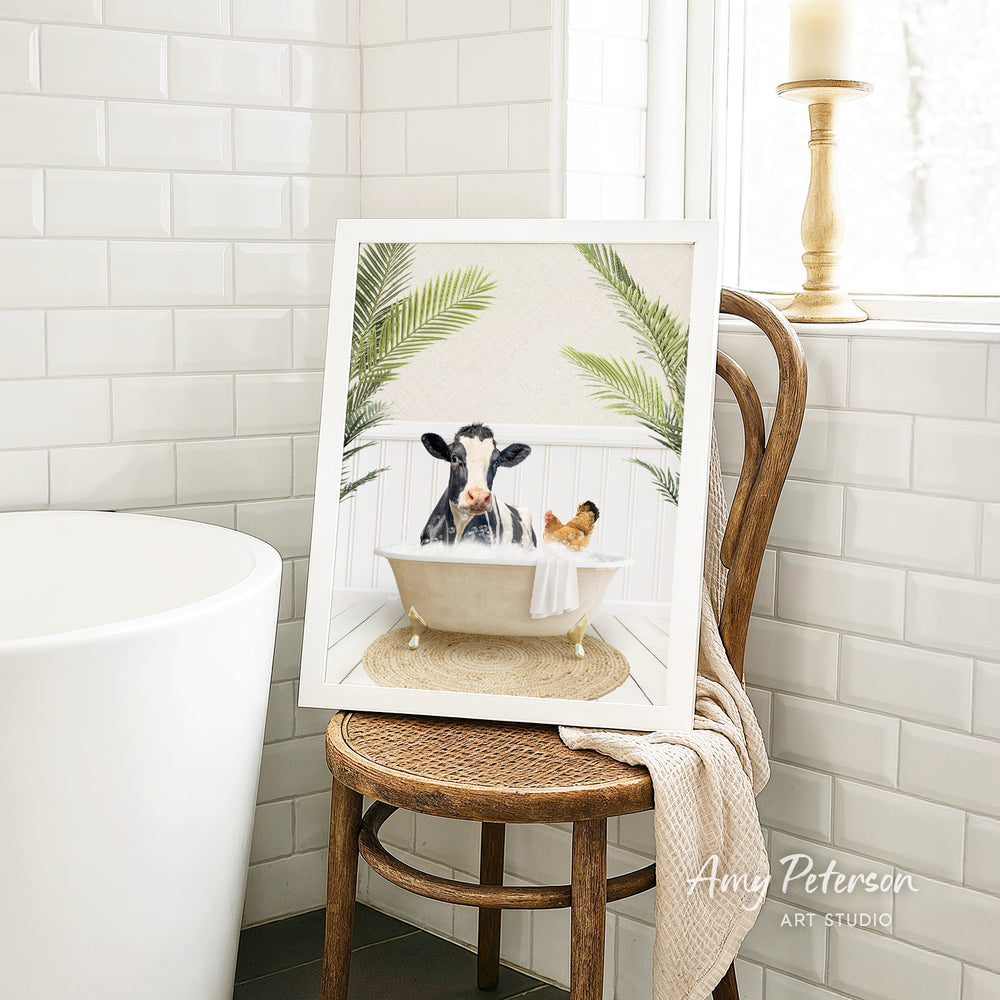 A framed image depicting a cow sitting in a bathtub, surrounded by palm leaves, with a small dog in the tub next to it.