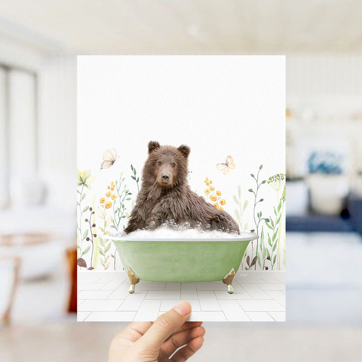 A hand holds up a framed image of a brown bear sitting in a green bathtub filled with bubbles, surrounded by a floral pattern.