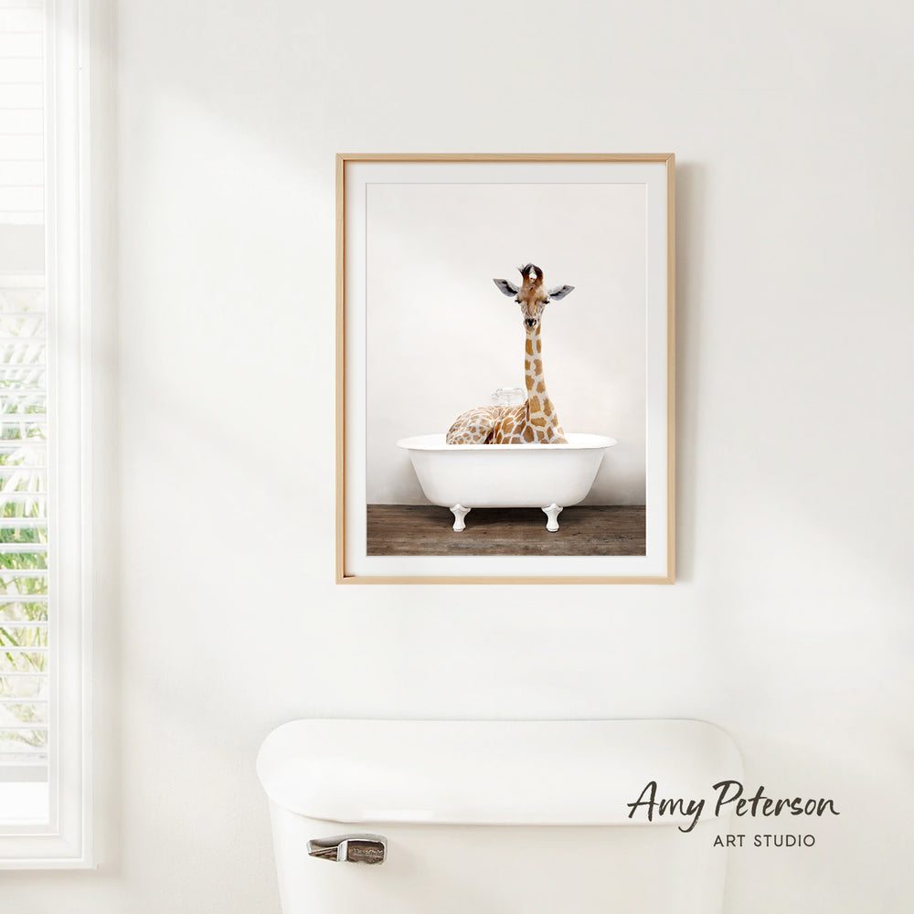 A framed image of a giraffe sitting in a white bathtub, with a window and a toilet visible in the background.