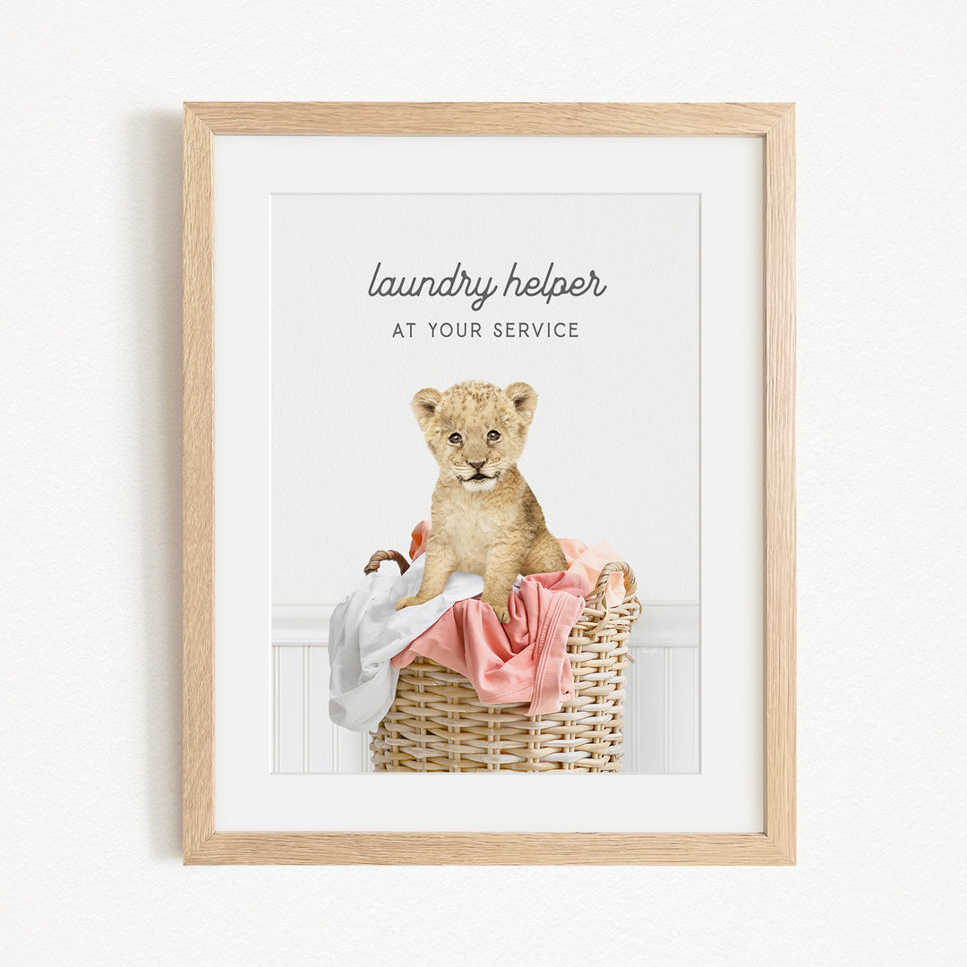 A framed image of a young lion cub sitting in a wicker basket filled with clothes, with the text "laundry helper at your service" written above the image.