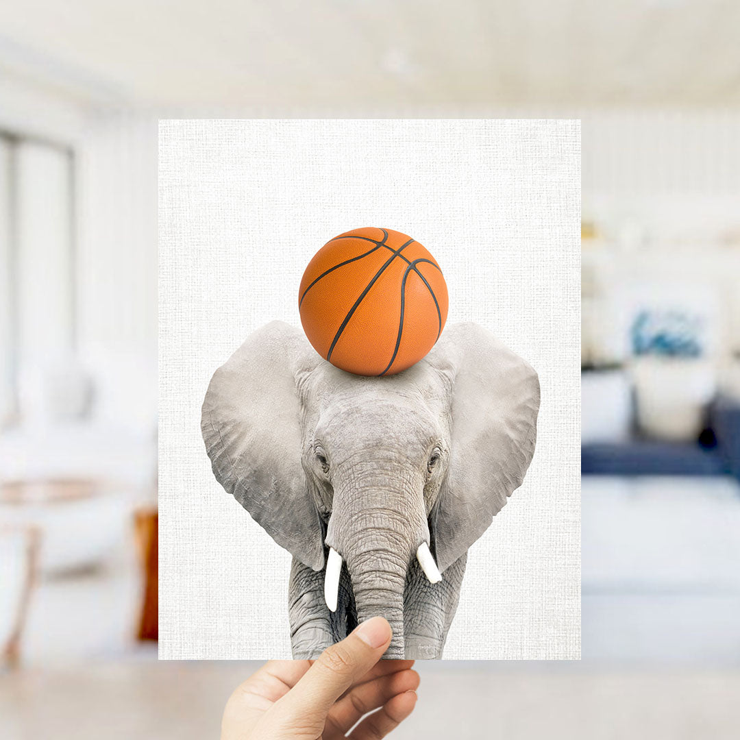 A hand holds up a framed image of a gray elephant with an orange basketball resting on its head.