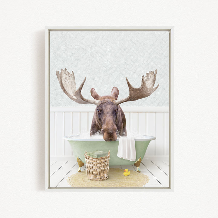 A moose with large antlers is sitting in a green bathtub filled with water, surrounded by a basket of rubber ducks and a towel.