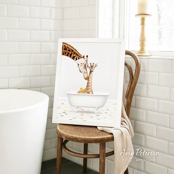 A framed image of two giraffes sitting in a bathtub, with one giraffe appearing to be bathing the other. The image is displayed on a wooden chair in a bathroom setting.