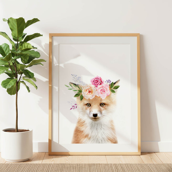A framed print of a fox wearing a floral crown is displayed on a white wall, with a potted plant to the left.