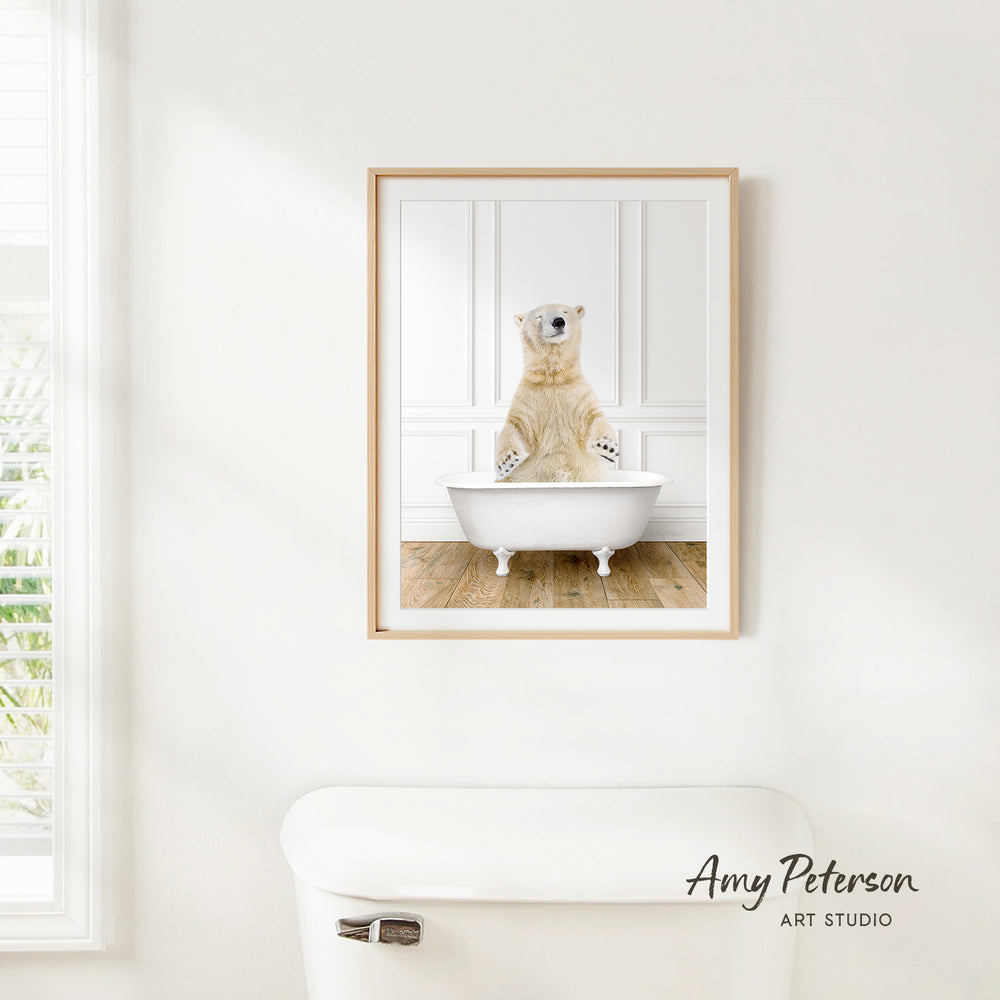 A polar bear sitting in a bathtub, as depicted in a framed artwork, is hanging on a white wall in a bathroom.