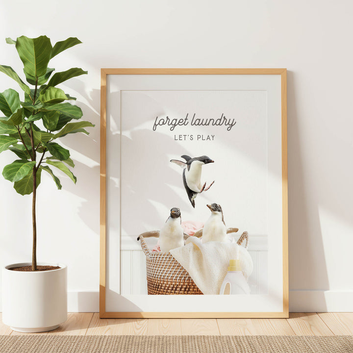 A framed poster with a playful penguin scene, featuring a basket of penguins and the text "Forget laundry, let's play".