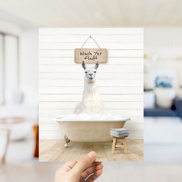 A hand holds up a framed image of a white llama sitting in a bathtub filled with bubbles, with a sign above it that says "Wash Your Fluff".