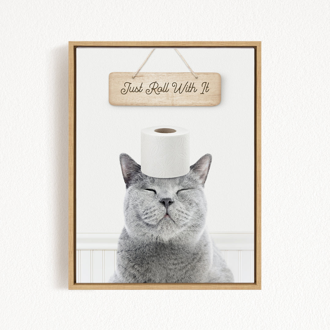 A gray cat with its eyes closed and a roll of toilet paper on its head, with a wooden sign above that says "Just Roll With It".