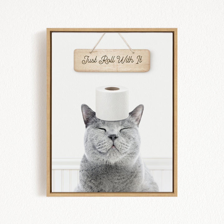 A gray cat with its eyes closed and a roll of toilet paper on its head, with a wooden sign above that says "Just Roll With It".