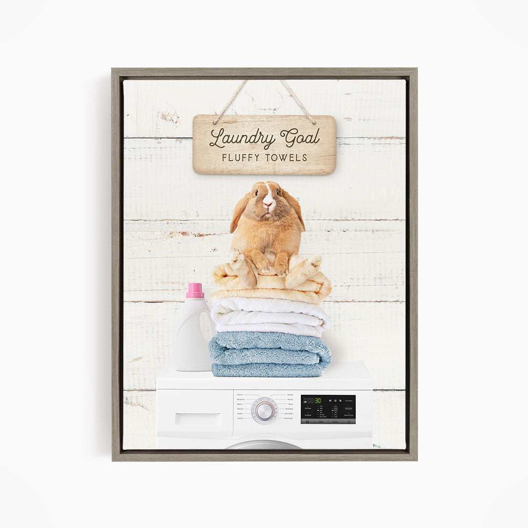 A wooden sign with the text "Laundry Goal Fluffy Towels" hangs above a stack of folded towels, with a stuffed rabbit sitting atop the towels.
