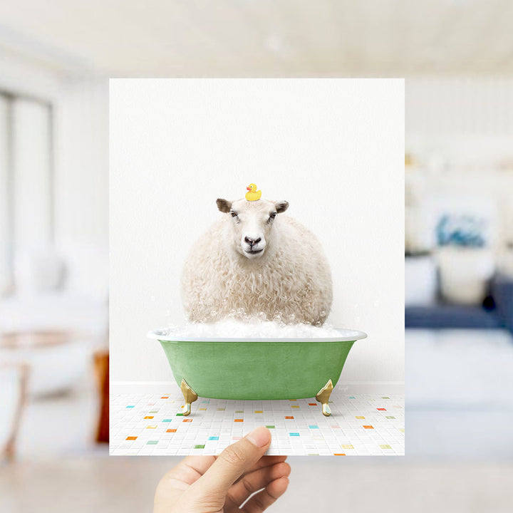 A hand holds up a framed image of a sheep sitting in a green bathtub filled with bubbles, with a yellow rubber duck perched on its head.