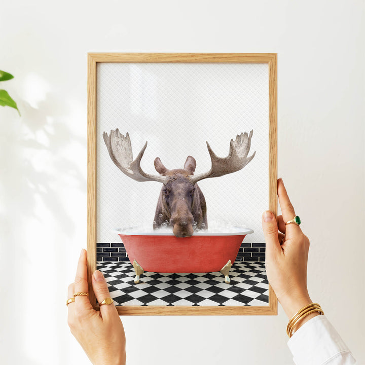 A person's hands are holding a framed picture of a moose in a bathtub, with a black and white checkered floor visible beneath the tub.
