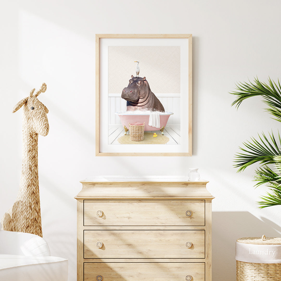 A framed illustration of a hippopotamus sitting in a pink bathtub, surrounded by a basket of flowers and a giraffe figurine, is displayed on a white wall above a wooden dresser.