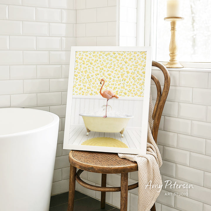 A framed painting of a pink flamingo sitting in a yellow bathtub is displayed on a wooden chair in a bathroom setting.