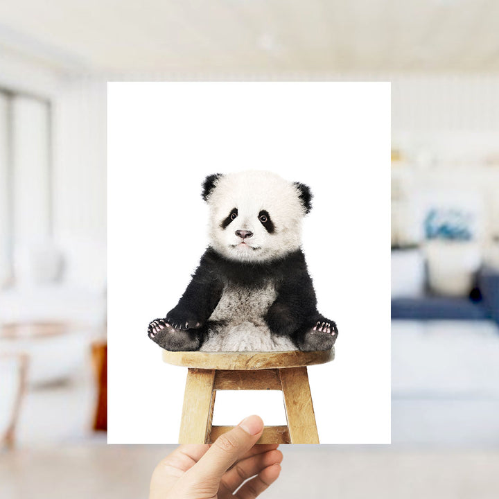 A hand is holding up a framed image of a panda bear sitting on a wooden stool.