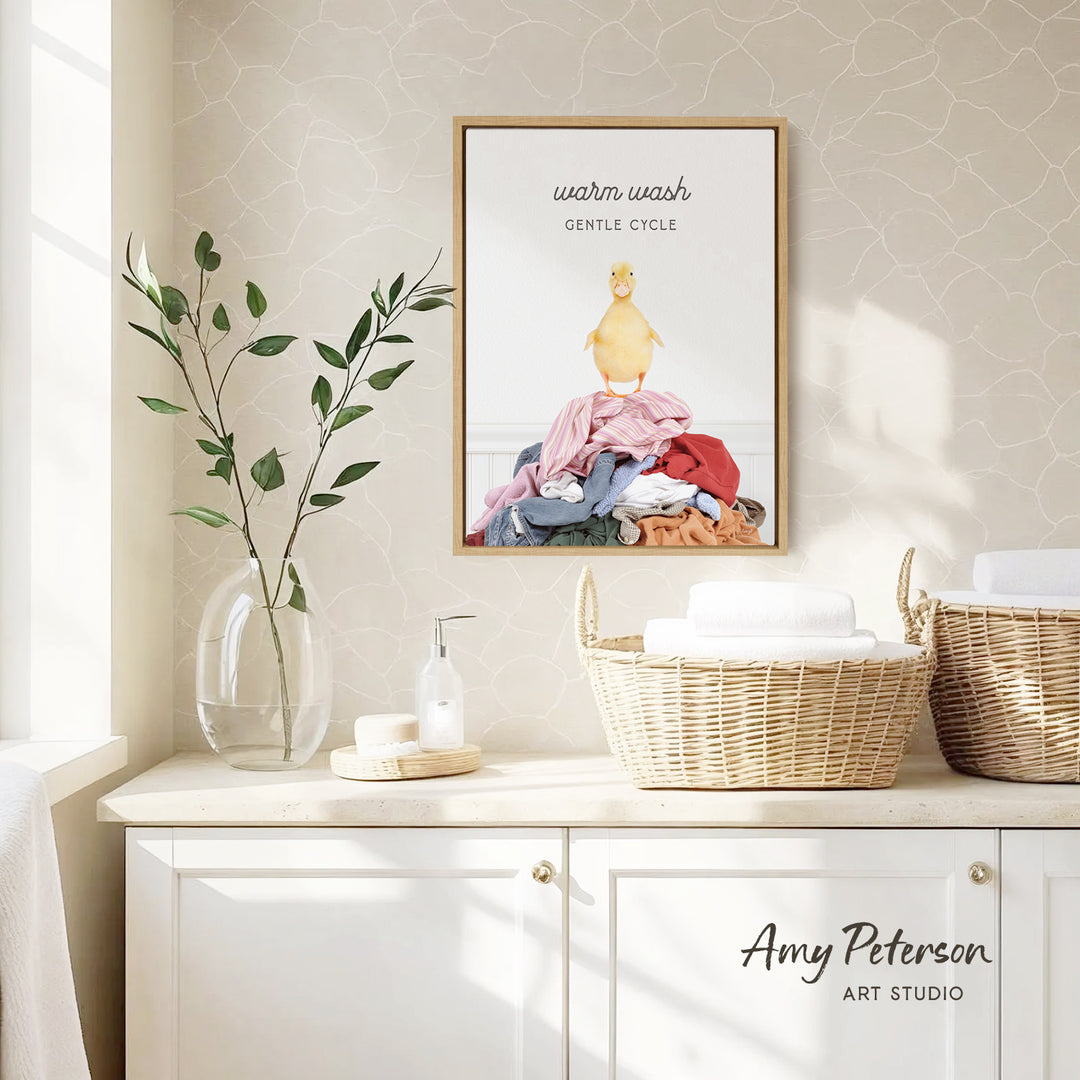 a bathroom counter with a framed poster of a duck sitting on a pile of laundry, a vase with green leaves, and baskets of towels.