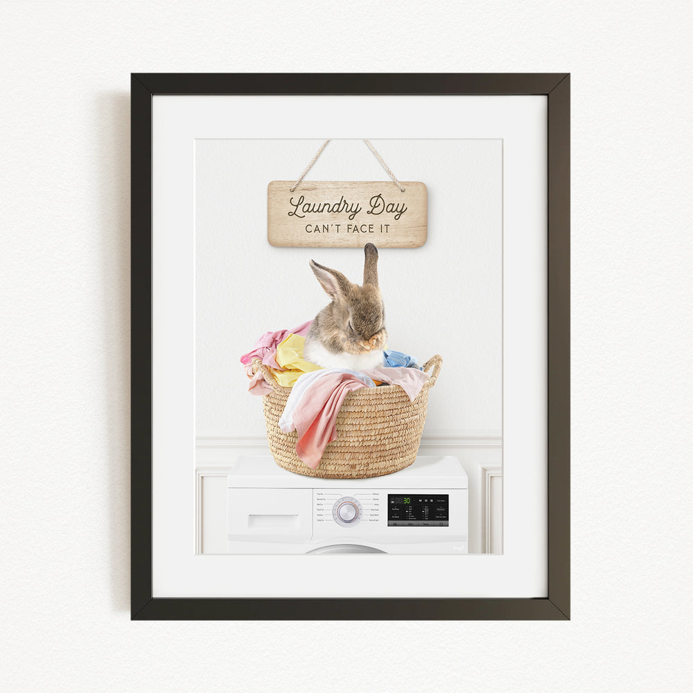 A framed image of a rabbit sitting in a basket filled with laundry, with a sign above that says "Laundry Day Can't Face It".