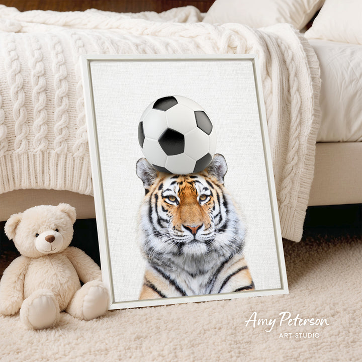 A framed print of a tiger's face with a soccer ball on its head, placed on a bed next to a teddy bear.