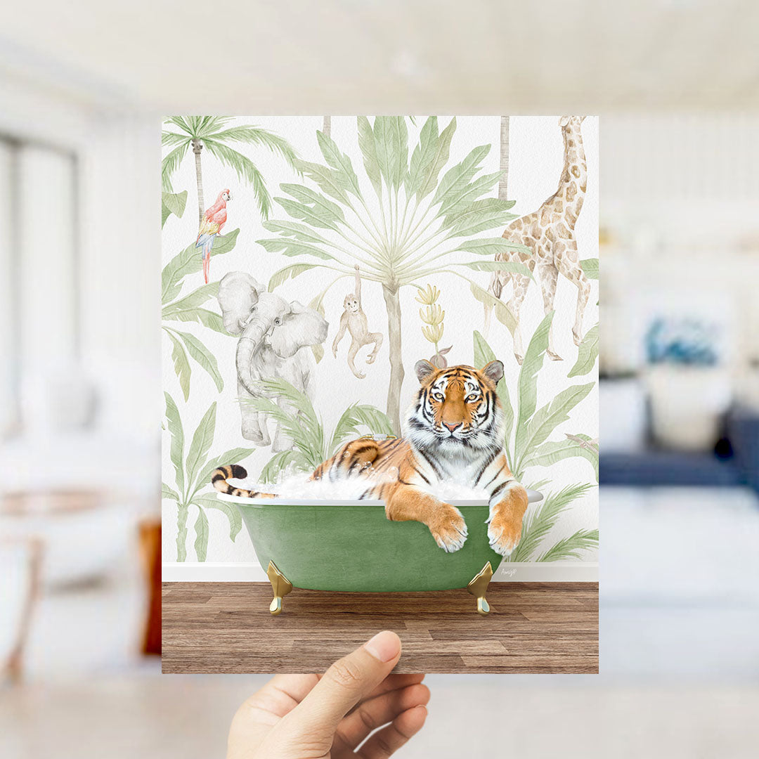 A hand holds up a framed print featuring a tiger lying in a bathtub surrounded by tropical foliage, including a giraffe, elephant, and parrot.
