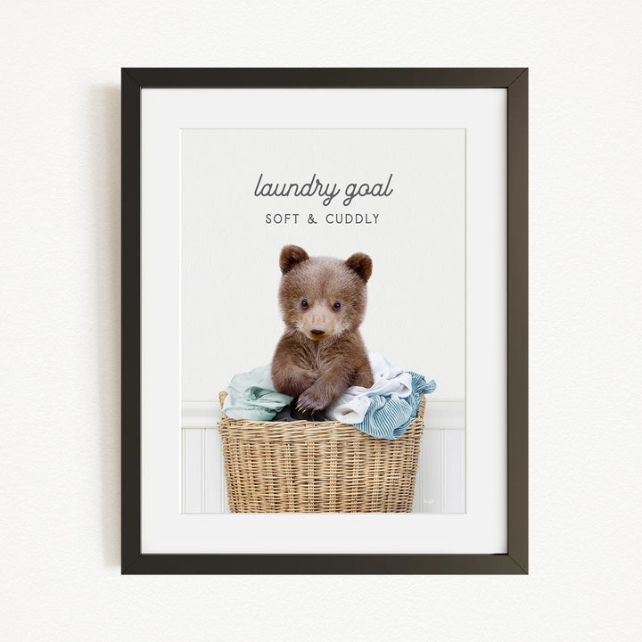 A cute brown bear sitting in a wicker laundry basket filled with clothes, with the text "laundry goal SOFT & Cuddly" displayed above the bear.