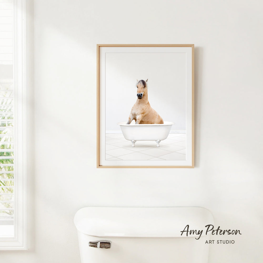 A framed image of a horse sitting in a bathtub is hanging on a white wall.