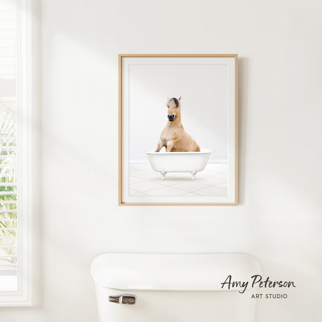 A framed image of a horse sitting in a bathtub is hanging on a white wall.