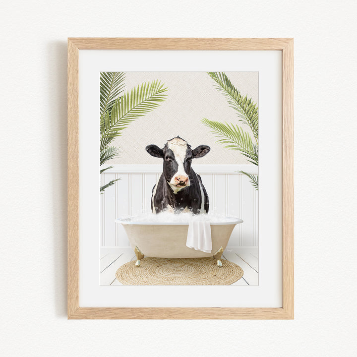 A black and white cow is sitting in a bathtub filled with water, surrounded by green palm leaves.