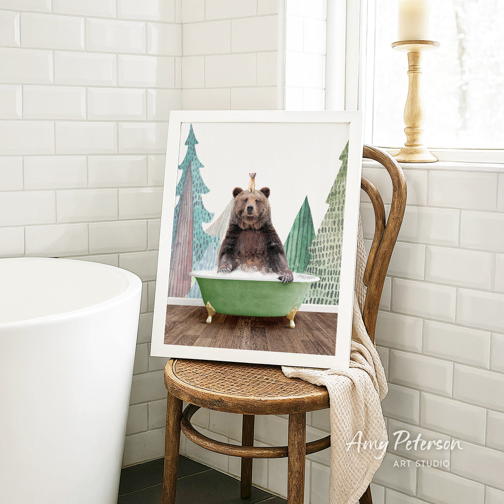 A framed painting of a bear sitting in a green bathtub, surrounded by trees, is displayed on a wooden chair in a bathroom setting.