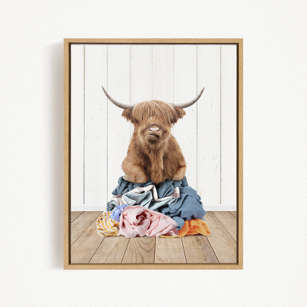 A brown cow with long horns is sitting atop a pile of clothes on a wooden floor.