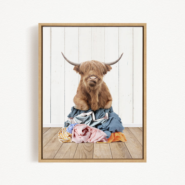 A brown cow with long horns is sitting atop a pile of clothes on a wooden floor.