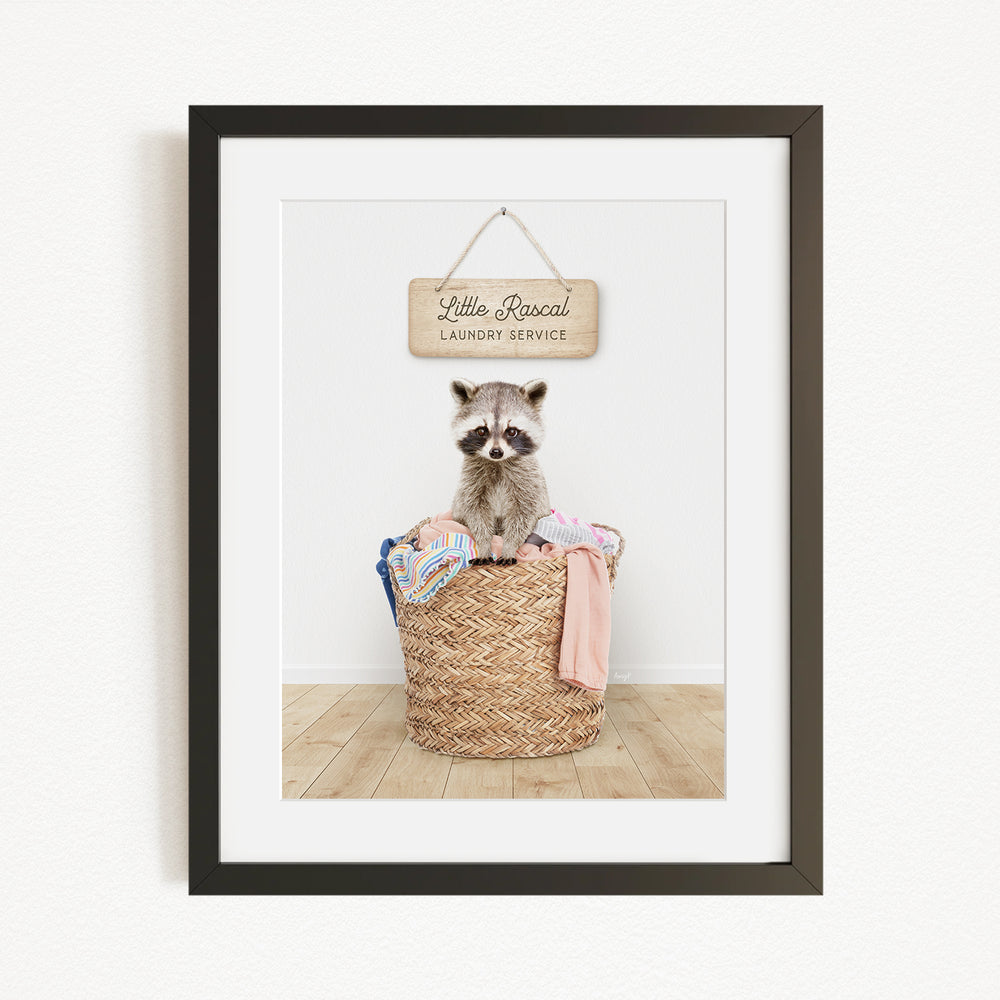 A raccoon sitting in a woven laundry basket, with a sign above that reads "Little Racquel Laundry Service".