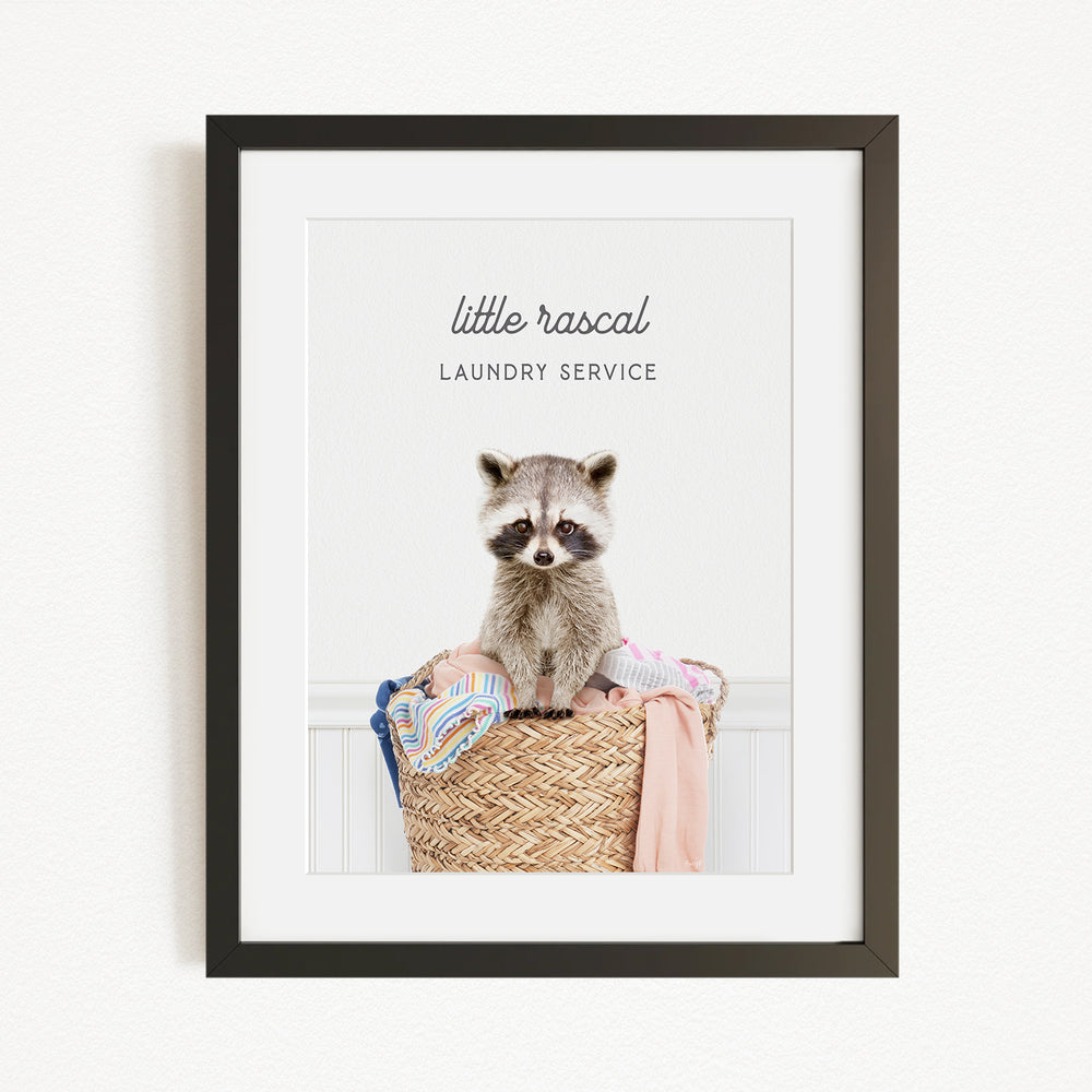 A raccoon sitting in a woven laundry basket, with the text "little rascal Laundry Service" displayed above the raccoon.