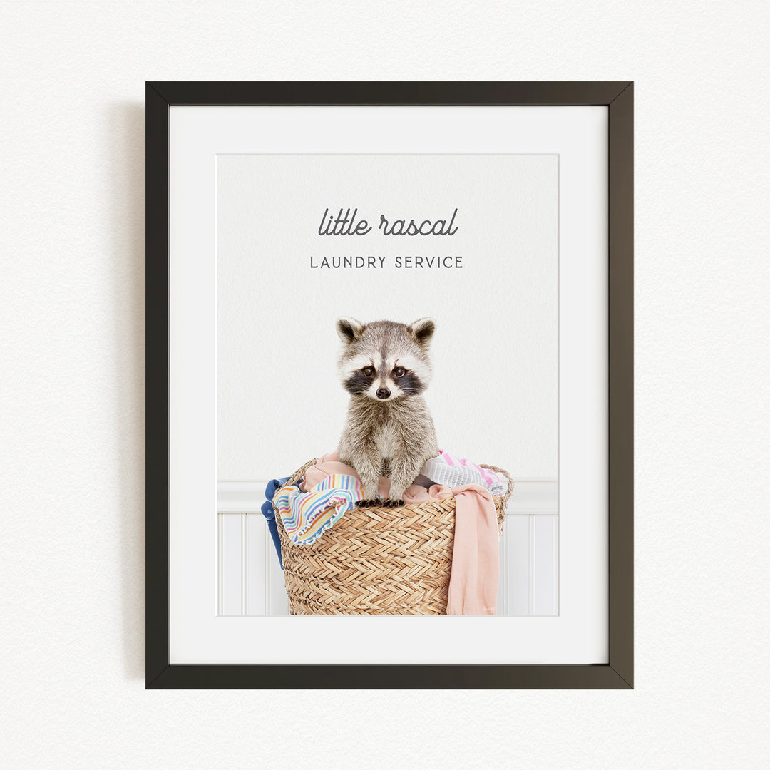 A raccoon sitting in a woven laundry basket, with the text "little rascal Laundry Service" displayed above the raccoon.