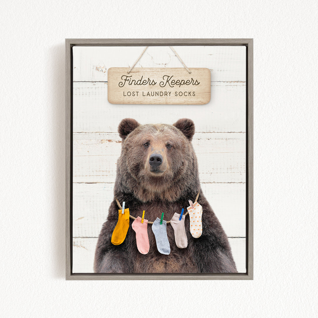 A framed image of a brown bear wearing colorful socks hanging on a clothesline, with a sign above that reads "Finders Keepers Lost Laundry Socks".