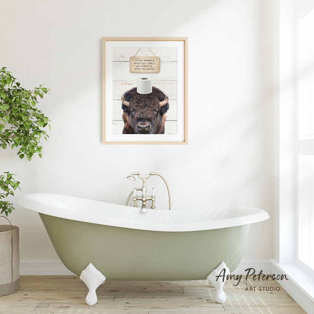 a bathroom with a green clawfoot bathtub, a framed picture of a bison with a sign above it, and a potted plant to the left.